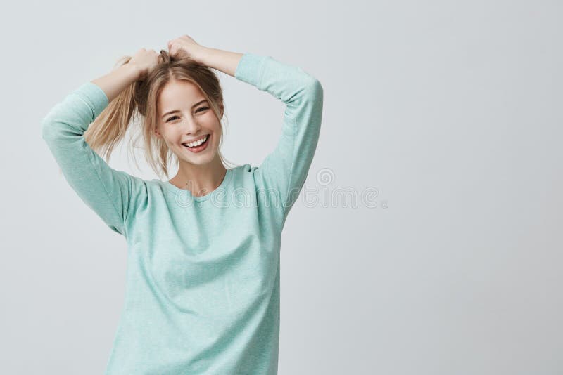 Human face expressions and emotions. Positive young beautiful female with dyed blonde straight hair in ponytail dressed in casual clothing looking at camera and pleasantly smiling, rejoicing her life. Human face expressions and emotions. Positive young beautiful female with dyed blonde straight hair in ponytail dressed in casual clothing looking at camera and pleasantly smiling, rejoicing her life