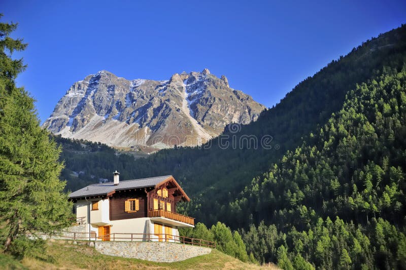 An alpine chalet in a forested alpine valley in Switzerland with a mountain peak beyond. Space for text in the clear blue sky. An alpine chalet in a forested alpine valley in Switzerland with a mountain peak beyond. Space for text in the clear blue sky.