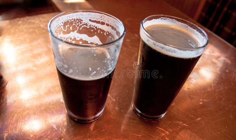 2 tall glasses of dark beer on a copper top table with 1 half empty. 2 tall glasses of dark beer on a copper top table with 1 half empty