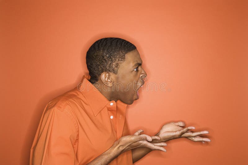 Side view of young African-American man on orange background expressing anger towards unseen person. Side view of young African-American man on orange background expressing anger towards unseen person.