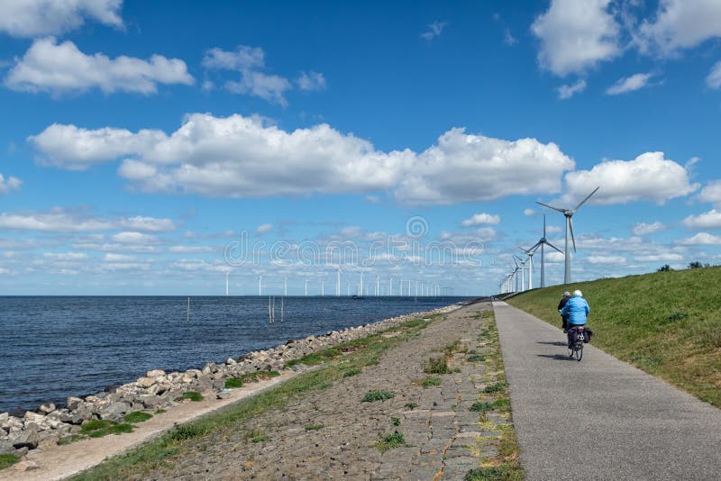 Dutch dike along IJsselmeer with two senior cyclists and wind turbines. Dutch dike along IJsselmeer with two senior cyclists and wind turbines