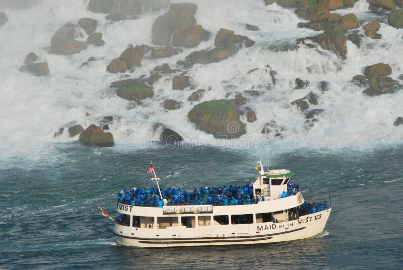 The boat Maid of the Mist sails below Niagara Falls, USA. Its upper and front deck packed with tourists with blue  ponchos. In the back the boulders of the lower part of the falls, lapped by sparkling waters. Hazy by the spraying waters. The boat Maid of the Mist sails below Niagara Falls, USA. Its upper and front deck packed with tourists with blue  ponchos. In the back the boulders of the lower part of the falls, lapped by sparkling waters. Hazy by the spraying waters.