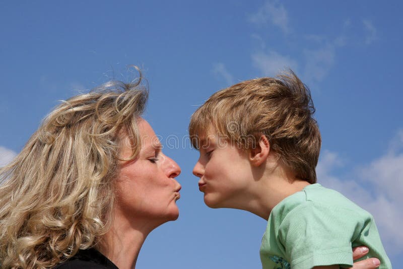 A beautiful mother giving a kiss to her cute 7-years old son photographed in the summer sun with blue sky and clouds in the background. A beautiful mother giving a kiss to her cute 7-years old son photographed in the summer sun with blue sky and clouds in the background