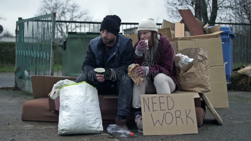 два грязных и плохо одетых бездомных человека мужчина и женщина сидят рядом с кучей мусора и едят и пьют что-нибудь