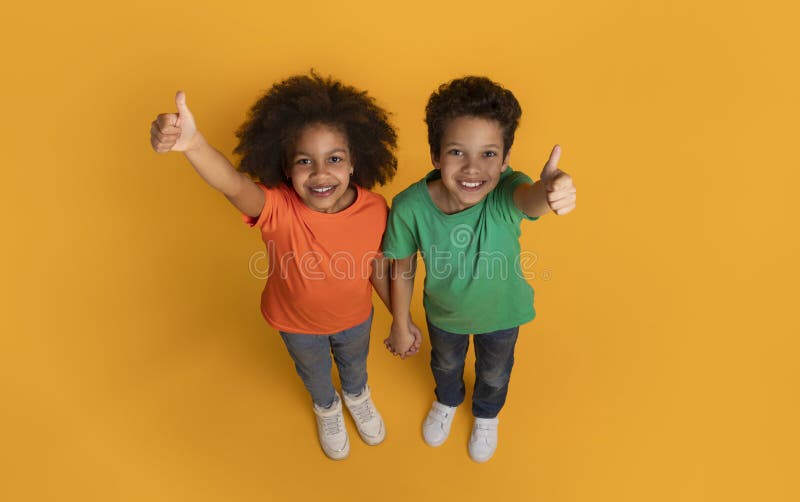 A cheerful African American boy and girl are standing on a bright yellow surface, looking upwards into the camera with joyful smiles, giving a thumbs up gesture, top view. A cheerful African American boy and girl are standing on a bright yellow surface, looking upwards into the camera with joyful smiles, giving a thumbs up gesture, top view