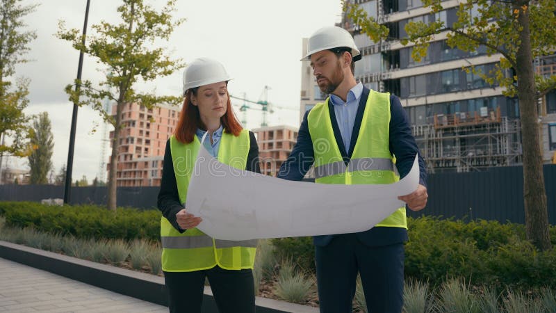 Two people workers builders in safety vests uniform hard hats discuss blueprint document paper plan of unfinished construction site discussion architects contractors men women talk in city civil build. High quality 4k footage. Two people workers builders in safety vests uniform hard hats discuss blueprint document paper plan of unfinished construction site discussion architects contractors men women talk in city civil build. High quality 4k footage