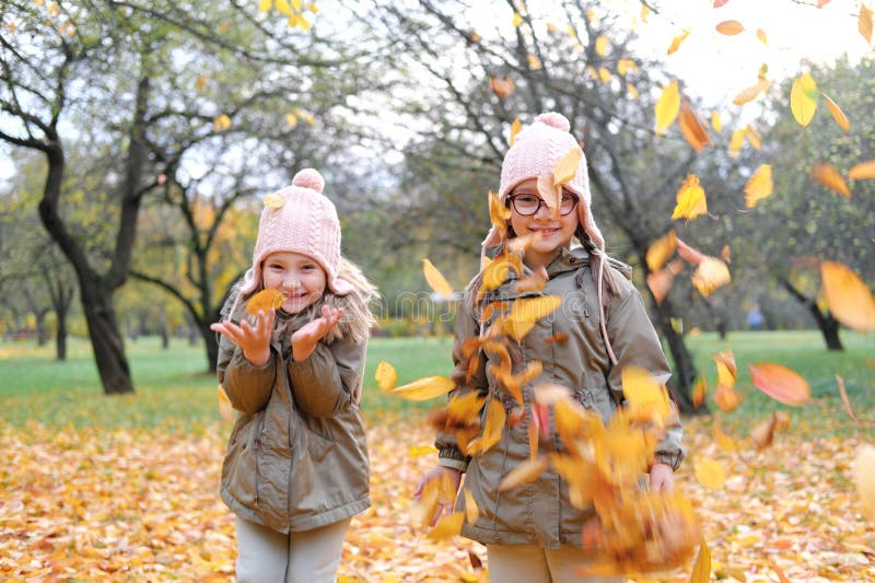 Two twin girls throw up a large armful of autumn leaves. Girls have fun in autumn park. Horizontal photo. Two twin girls throw up a large armful of autumn leaves. Girls have fun in autumn park. Horizontal photo