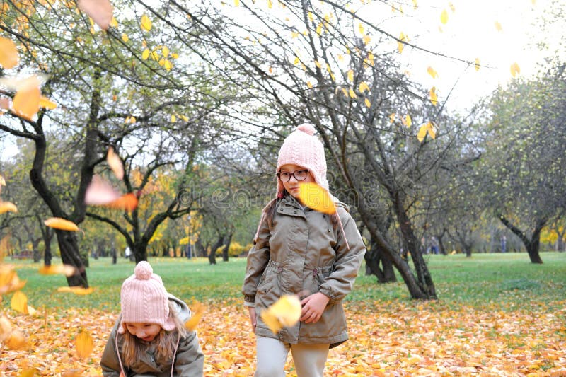 Two twin girls collect autumn leaves to throw them up. Girls have fun in autumn park. Horizontal photo. Two twin girls collect autumn leaves to throw them up. Girls have fun in autumn park. Horizontal photo