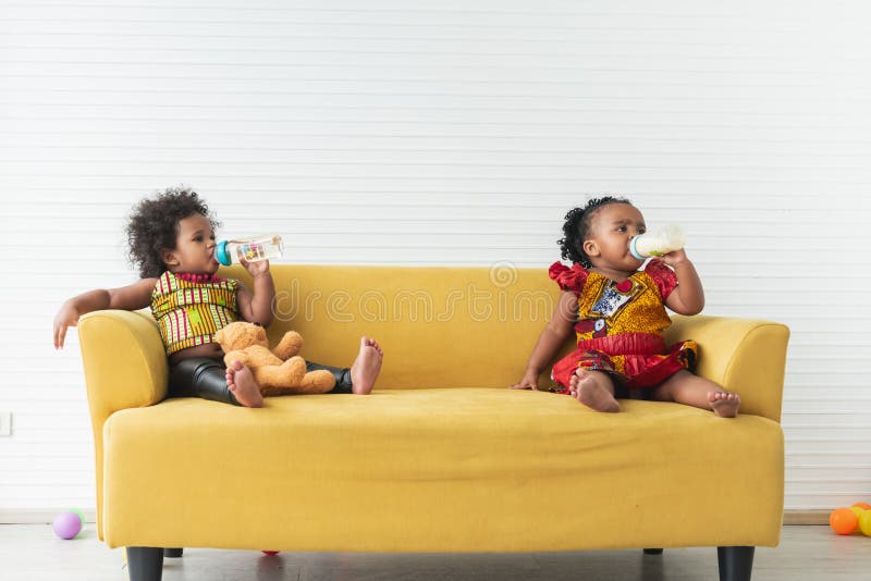 Two Nigerian baby girls, aged 2 and 1, sitting on a yellow sofa, drinking milk and water from bottles. on white background. to African family and baby food concept. Two Nigerian baby girls, aged 2 and 1, sitting on a yellow sofa, drinking milk and water from bottles. on white background. to African family and baby food concept