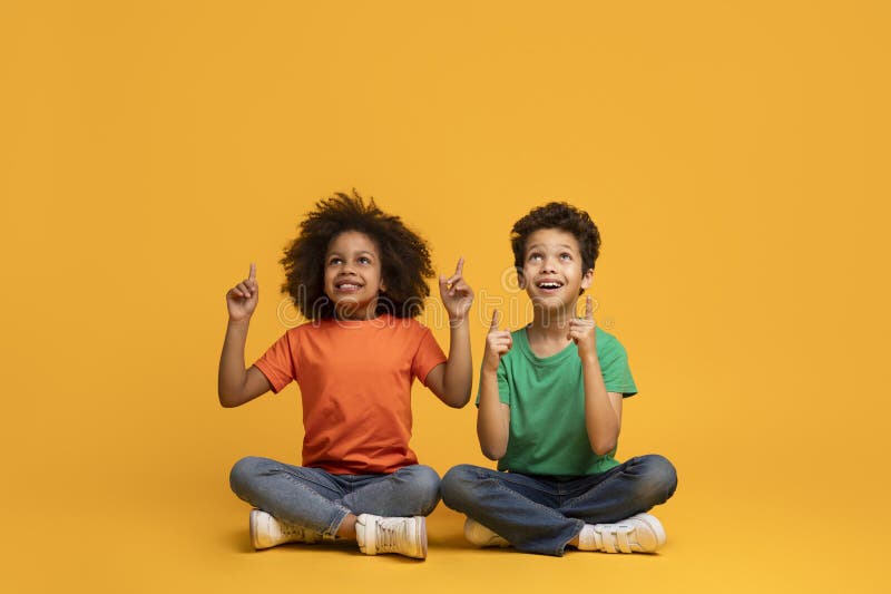 Two African American children, a boy and a girl, are sitting cross-legged on the floor with their fingers extended upwards, yellow background. Two African American children, a boy and a girl, are sitting cross-legged on the floor with their fingers extended upwards, yellow background