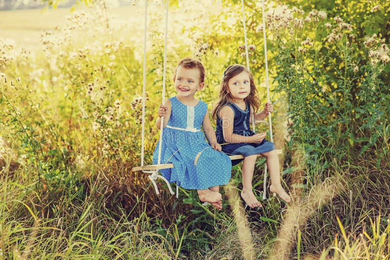 Two little girls in blue dress on swing in summertime on field. Happy children playing outdoors in sunset in summer. Cheerful and active barefoot kids close to nature. Dangerous harving mites on grass. Two little girls in blue dress on swing in summertime on field. Happy children playing outdoors in sunset in summer. Cheerful and active barefoot kids close to nature. Dangerous harving mites on grass