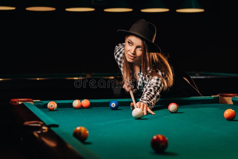 A girl in a hat in a billiard club with a cue in her hands hits a ball.Playing billiards. A girl in a hat in a billiard club with a cue in her hands hits a ball.Playing billiards.