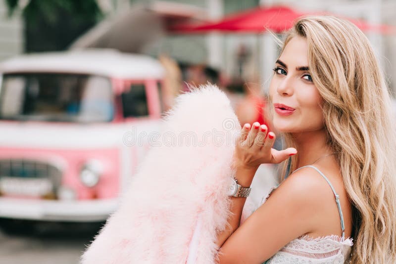 Closeup portrait attractive girl holding pink fur stole in hand on retro car background. She has long blonde hair, bare shoulder, sending a kiss to camera. Closeup portrait attractive girl holding pink fur stole in hand on retro car background. She has long blonde hair, bare shoulder, sending a kiss to camera