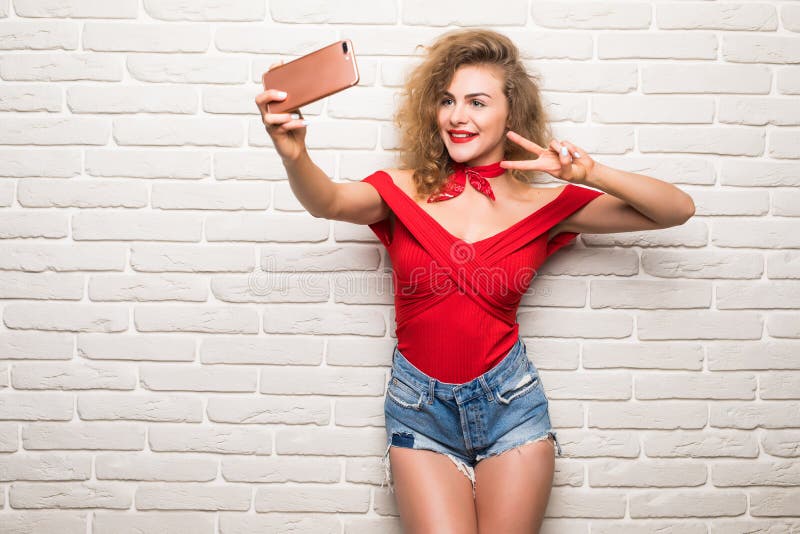 Девушка принимая selfie умную камеру телефона возбудила счастливую женщину улыбки над белой стеной офиса кирпича стоковое фото rf