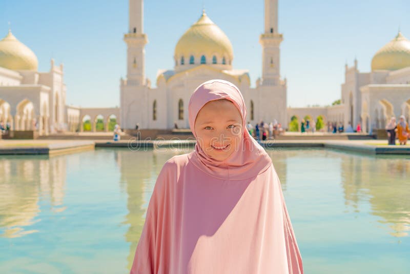 Девушка ребенк в розовом hijab сидит рядом с белой мечетью и улыбками r стоковое фото rf Девушка ребенк в розовом hijab сидит рядом с белой мечетью и улыбками r стоковое фото rf