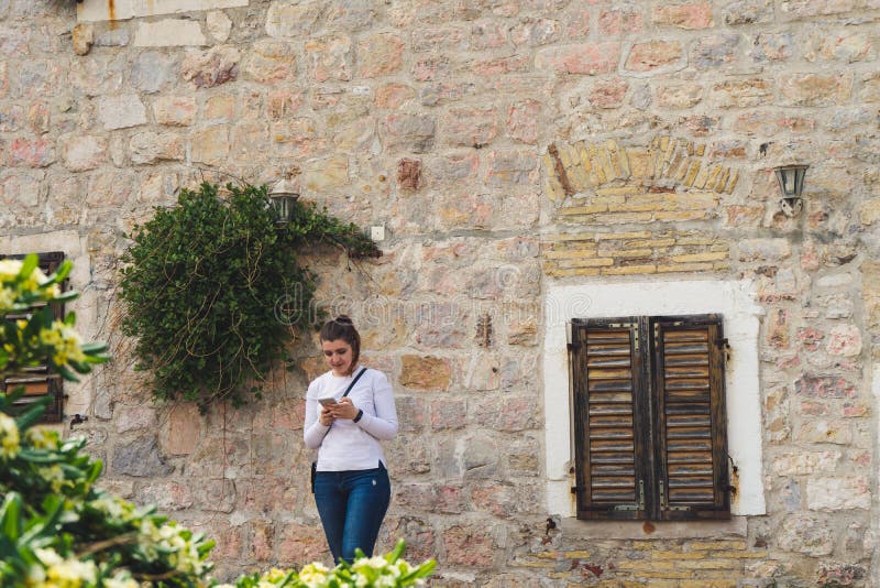 Girl with white long shirt sending a message in smart phone and stone house with flowers in the facade. Woman typing in cell phone and wood windows in mediterranean town, texting, wall, conversation, traveler, people, smartphone, traditional, mobile, cellphone, lady, female, sms, chat, using, tourist, communication, online, technology, wooden. Girl with white long shirt sending a message in smart phone and stone house with flowers in the facade. Woman typing in cell phone and wood windows in mediterranean town, texting, wall, conversation, traveler, people, smartphone, traditional, mobile, cellphone, lady, female, sms, chat, using, tourist, communication, online, technology, wooden