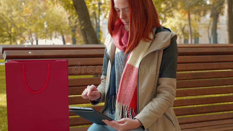 Девушка с лисиными волосами, сидящая на скамейке на солнечной погоде, пользуется планшетом и делает онлайн заказ