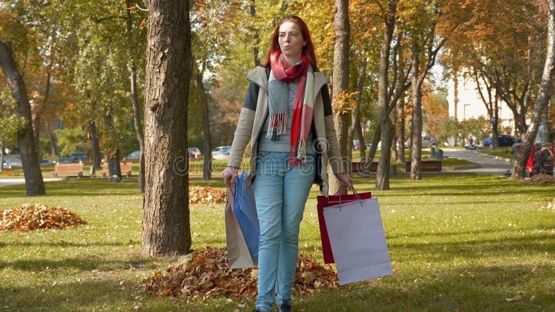 Девушка с лисистой шерстью ходит по парку с покупками в полиэтиленовых пакетах
