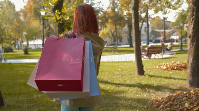 Девушка с лисистой шерстью ходит по парку с покупками в полиэтиленовых пакетах