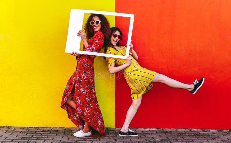 Two women friends with blank photo frame standing against colored wall outdoors. Female travelers posing at camera with empty picture frame. Two women friends with blank photo frame standing against colored wall outdoors. Female travelers posing at camera with empty picture frame