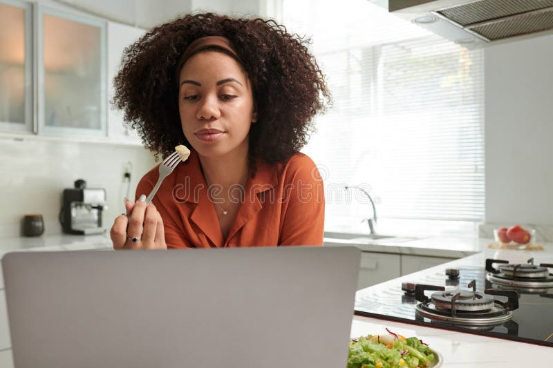 Businesswoman eating salad at kitchen counter and reading e-mails or articles on laptop screen. Businesswoman eating salad at kitchen counter and reading e-mails or articles on laptop screen