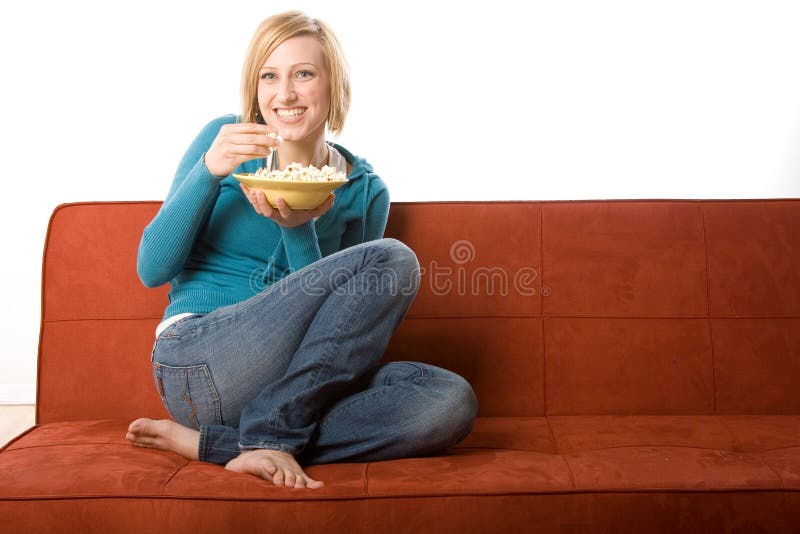 Young adult blond female model sits on orange colored couch eating popcorn from a bowl. Young adult blond female model sits on orange colored couch eating popcorn from a bowl.