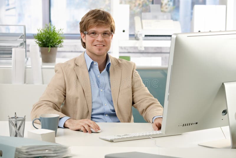 Young businessman sitting at office desk, using computer, looking at camera, smiling. Young businessman sitting at office desk, using computer, looking at camera, smiling.