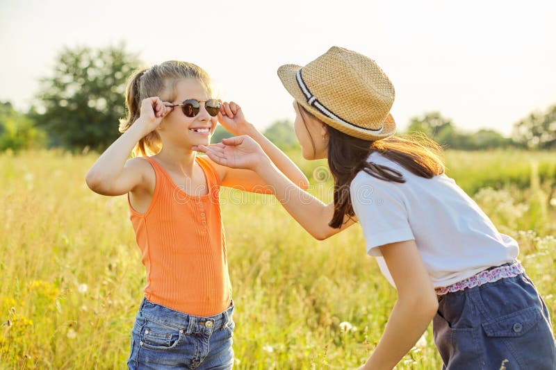 Children are having fun in nature. Two girls laugh, look at themselves in the mirror of sunglasses, sunny summer meadow with grasses background. Childhood, fun, happiness, summer concept. Children are having fun in nature. Two girls laugh, look at themselves in the mirror of sunglasses, sunny summer meadow with grasses background. Childhood, fun, happiness, summer concept