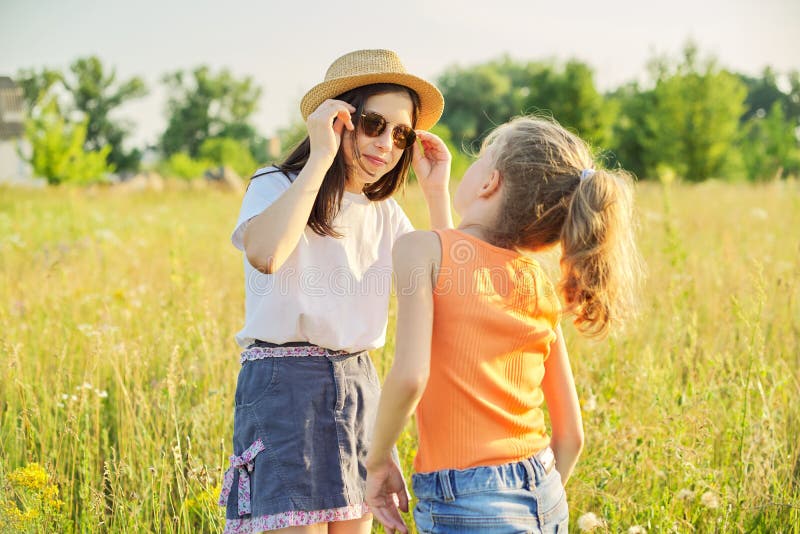 Children are having fun in nature. Two girls laugh, look at themselves in the mirror of sunglasses, sunny summer meadow with grasses background. Childhood, fun, happiness, summer concept. Children are having fun in nature. Two girls laugh, look at themselves in the mirror of sunglasses, sunny summer meadow with grasses background. Childhood, fun, happiness, summer concept