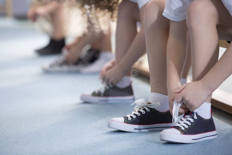 Close-up on school kids` legs while they`re sitting on a bench and tying their sport shoes for physical education activities. Close-up on school kids` legs while they`re sitting on a bench and tying their sport shoes for physical education activities