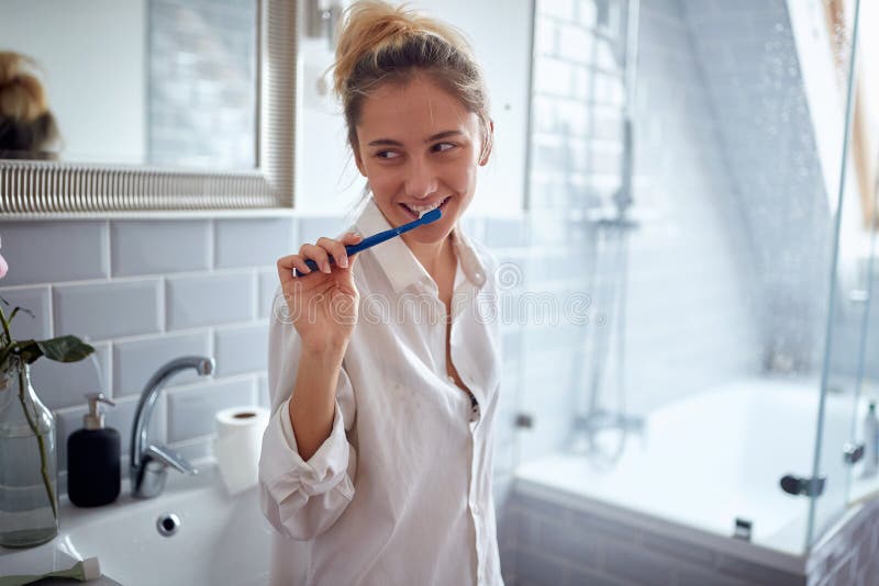 Satisfied young adult caucasian blonde female thinking with smile while brushing her teeth in the bathroom. Satisfied young adult caucasian blonde female thinking with smile while brushing her teeth in the bathroom