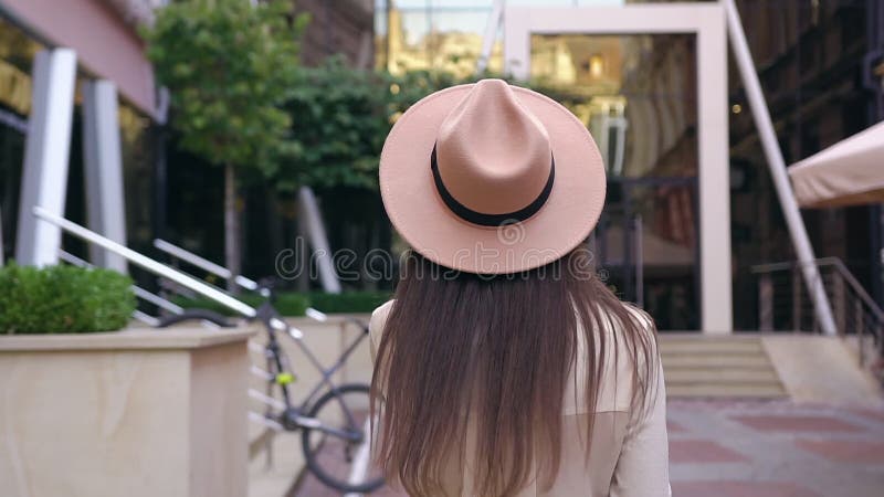 Довольно радостная 30-летняя женщина в модисной шляпе разворачивается, ходя рядом с красивым современным зданием и разговаривая о