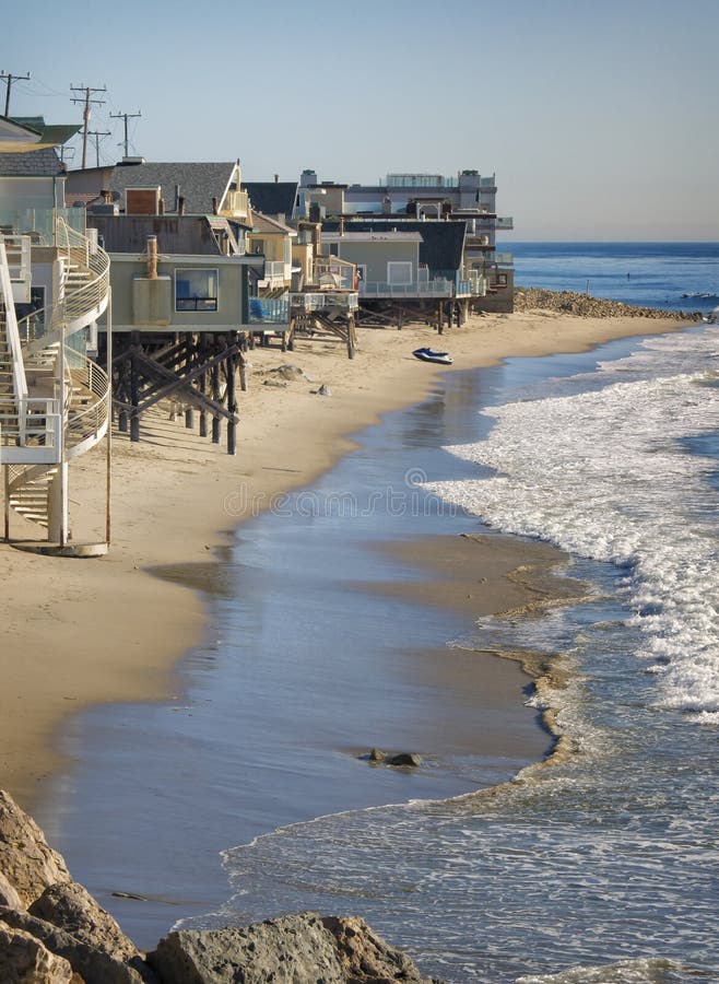 Houses on stilts sit close to waters edge on the sand of the beach with surfers in the distance on the coast of the Pacific Ocean, just north of Malibu, a popular and famous beach front area in southern California, just south of Los Angeles, frequented by tourists and locals and known for movie stars who live in some of the coastal homes. Houses on stilts sit close to waters edge on the sand of the beach with surfers in the distance on the coast of the Pacific Ocean, just north of Malibu, a popular and famous beach front area in southern California, just south of Los Angeles, frequented by tourists and locals and known for movie stars who live in some of the coastal homes.