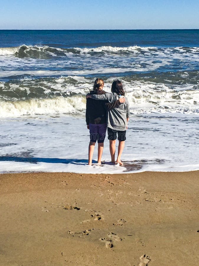 Two friends, cousins arm and arm standing in the waves gazing out into the ocean. Footprints in the sand walking to the water. Hugs from far away loved ones. Girls wearing sweatshirts and their pants rolled up so they can enjoy the Outer Banks of North Carolina shore. Large waves crashing on the beach. The vastness of the salt water of the Atlantic Ocean. Two friends, cousins arm and arm standing in the waves gazing out into the ocean. Footprints in the sand walking to the water. Hugs from far away loved ones. Girls wearing sweatshirts and their pants rolled up so they can enjoy the Outer Banks of North Carolina shore. Large waves crashing on the beach. The vastness of the salt water of the Atlantic Ocean