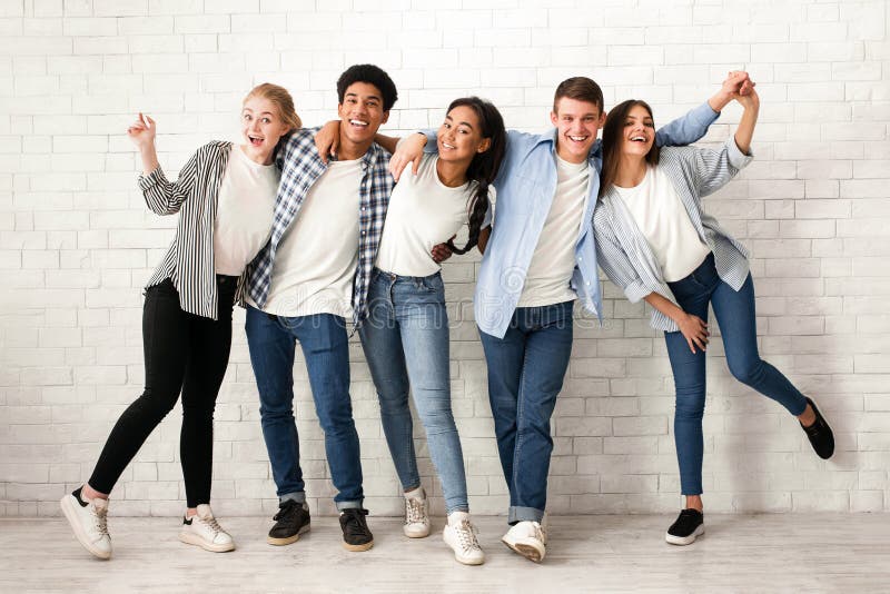 A group of five multiethnic teenagers are standing in a row against a white brick wall, smiling and laughing. They are dressed in casual clothing. A group of five multiethnic teenagers are standing in a row against a white brick wall, smiling and laughing. They are dressed in casual clothing