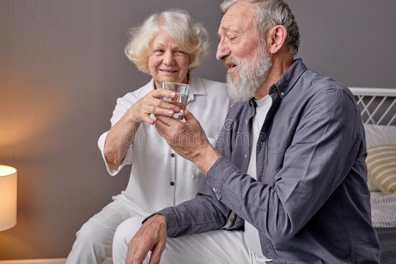 friednly pleasant wife give glass of water to retired husband, help and support, sitting on bed at home. in light room interior. friednly pleasant wife give glass of water to retired husband, help and support, sitting on bed at home. in light room interior