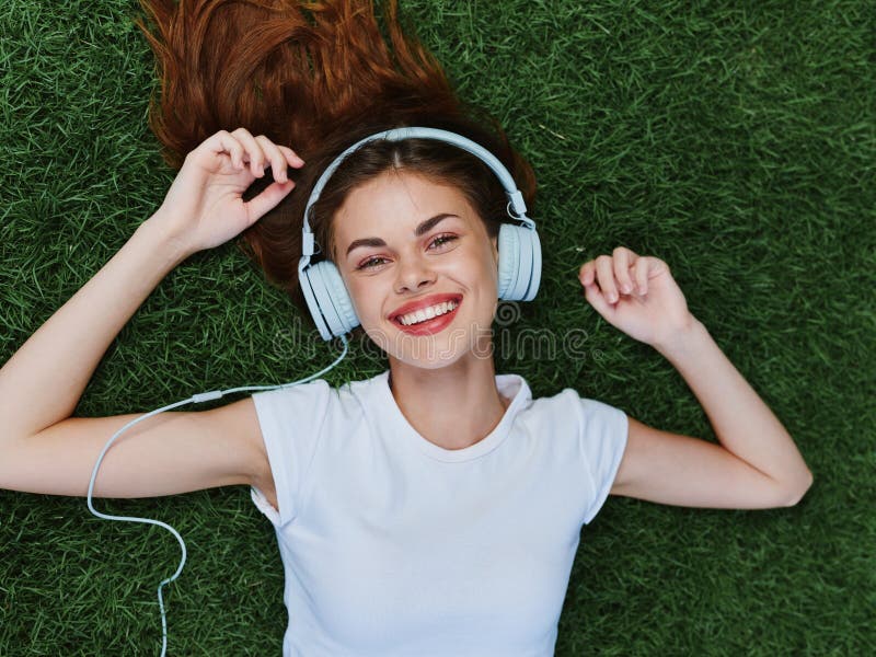 Woman portrait lying on the green grass on a sunny summer day listening to music in headphones in a. Woman portrait lying on the green grass on a sunny summer day listening to music in headphones in a