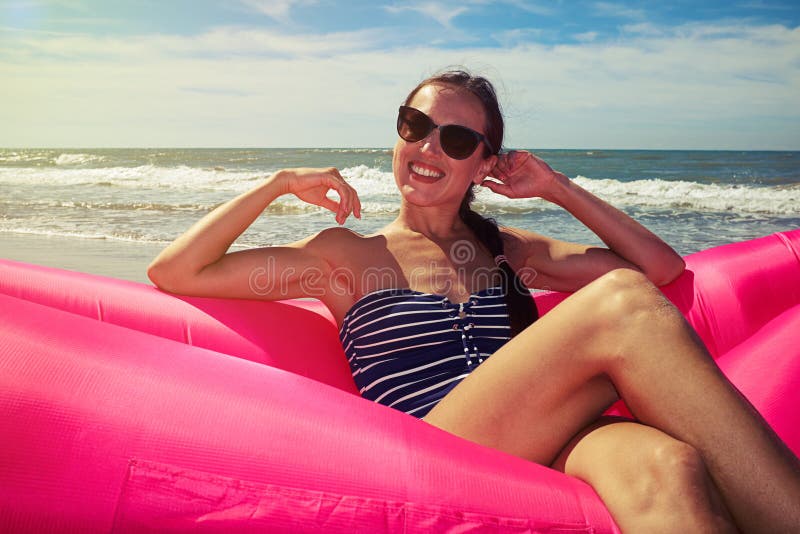 A pretty woman demonstrating her sincere smile, wearing brown sunglasses and blue and white striped swimming suit with the hands near her face reclining on a rosy airbed on a sunny day with the sky covered with some clouds. A pretty woman demonstrating her sincere smile, wearing brown sunglasses and blue and white striped swimming suit with the hands near her face reclining on a rosy airbed on a sunny day with the sky covered with some clouds