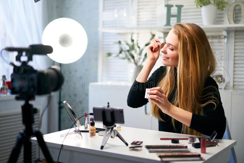 Beauty blogger woman filming daily makeup routine tutorial near camera on tripod. Influencer girl live streaming cosmetics product review in home studio. Vlogger female applies powder with brush. Beauty blogger woman filming daily makeup routine tutorial near camera on tripod. Influencer girl live streaming cosmetics product review in home studio. Vlogger female applies powder with brush.