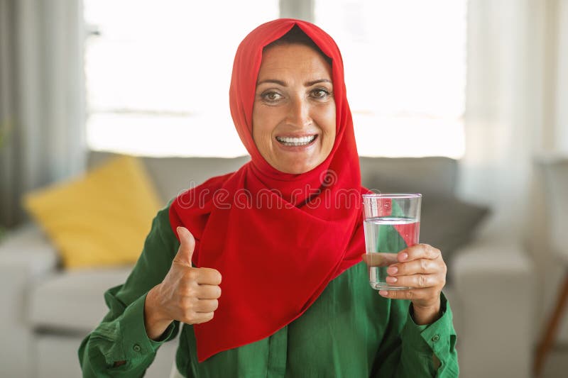 A woman wearing a red headscarf holds a glass filled with water. She is standing in an indoor setting, with a neutral background. Lady face is not shown, focusing on her hands and the glass of water. A woman wearing a red headscarf holds a glass filled with water. She is standing in an indoor setting, with a neutral background. Lady face is not shown, focusing on her hands and the glass of water.