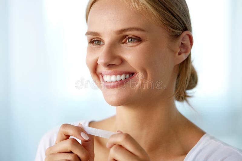 White Smile. Beautiful Smiling Woman With Healthy White Teeth Applying Teeth Whitening Strip. Portrait Of Happy Girl Using Dental Whitener. Dental Beauty, Tooth Care Concept. High Resolution Image. White Smile. Beautiful Smiling Woman With Healthy White Teeth Applying Teeth Whitening Strip. Portrait Of Happy Girl Using Dental Whitener. Dental Beauty, Tooth Care Concept. High Resolution Image