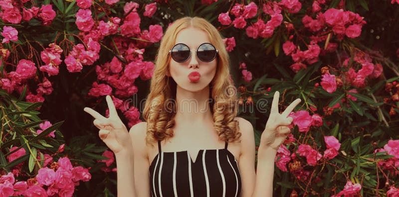 Portrait of beautiful woman blowing her red lips sending sweet air kiss on a pink roses flowers background. Portrait of beautiful woman blowing her red lips sending sweet air kiss on a pink roses flowers background