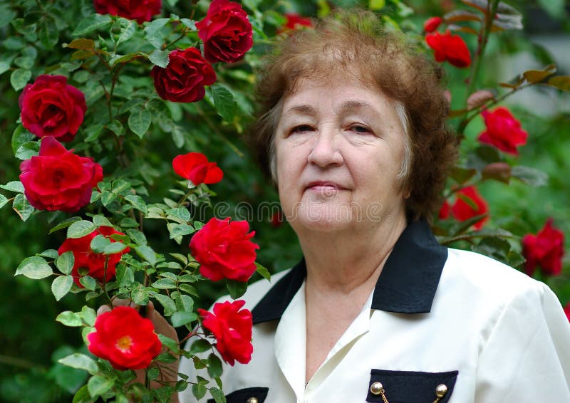 Elderly woman costs in the garden near to a bush of beautiful red roses. Elderly woman costs in the garden near to a bush of beautiful red roses.