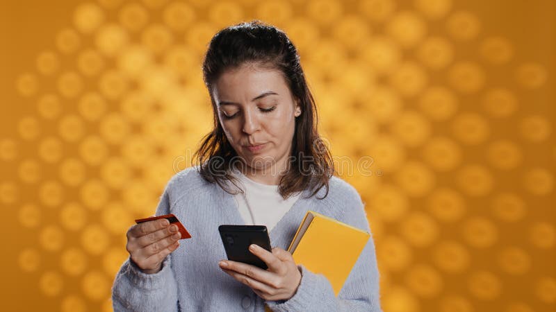 Woman holding books, purchasing more with phone, conveying joy of reading concept, studio background. Person buying novels online with credit card, showing appreciation for literature, camera B. Woman holding books, purchasing more with phone, conveying joy of reading concept, studio background. Person buying novels online with credit card, showing appreciation for literature, camera B