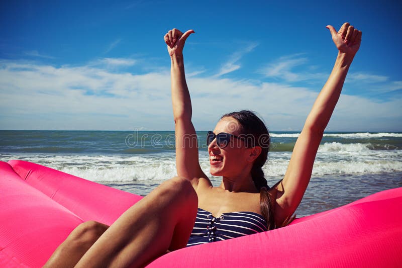 A female sitting on a deep-pink airbed on the beach holding her thumbs up and smiling. She’s wearing a striped swimming suit and dark sunglasses, her hair is put in a braid. A sunny day, blue sky and raging sea. A female sitting on a deep-pink airbed on the beach holding her thumbs up and smiling. She’s wearing a striped swimming suit and dark sunglasses, her hair is put in a braid. A sunny day, blue sky and raging sea