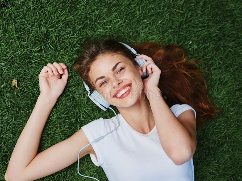 Woman listening to music with headphones in a white T-shirt on the green grass of the park lawn. Woman listening to music with headphones in a white T-shirt on the green grass of the park lawn