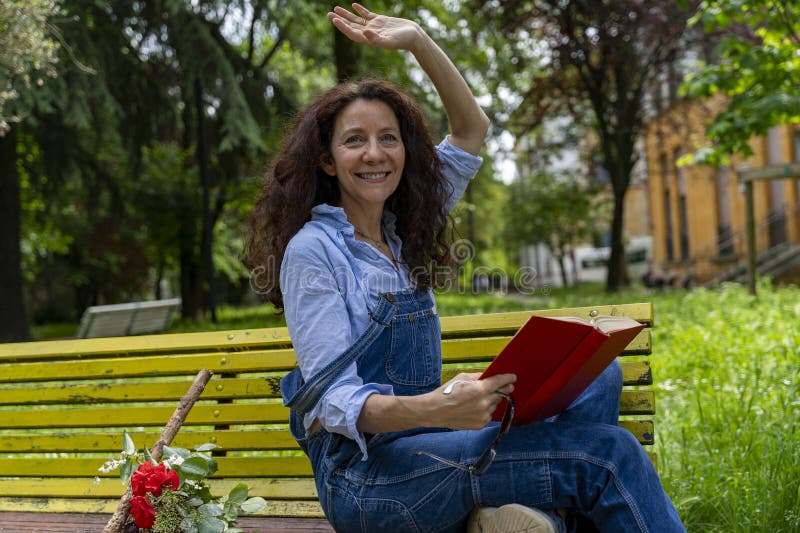 женщина средних лет, сидящая на жёлтой скамье, машет рукой, читая книгу стоковая фотография