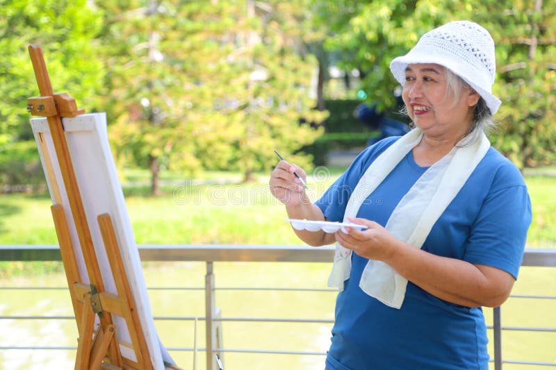 Женщина-старший рисует на свежем воздухе на Easel Mature Artist Creating Art in Nature Asian Lady Enjoing Outdoor Painting Hobby стоковые фотографии rf