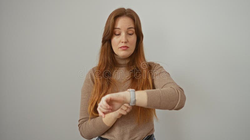 Женщина, указывающая на наручные часы на запястье, на белом фоне молодая рыжая женщина выглядит внимательной в коричневом акции видеоматериалы