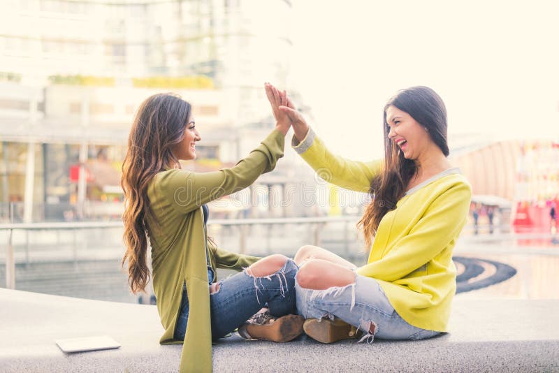 Two beautiful young women giving high five - Pretty girls sitting on a bench outdoors and having fun - Best girlfriends making a promise. Two beautiful young women giving high five - Pretty girls sitting on a bench outdoors and having fun - Best girlfriends making a promise